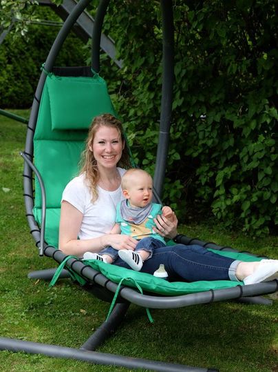 A woman is sitting on a green swing, holding a small child on her lap. The background is decorated with trees and grass.