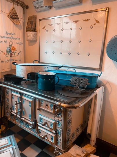 Eine alte Küche mit einem nostalgischen Kochofen und verschiedenen Kochutensilien. Die Wände sind mit dekorativen Elementen und Mustern geschmückt.
