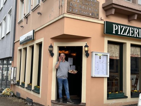 Un homme se tient devant une pizzeria et tient un plateau de plats à la main. Le bâtiment est accueillant et possède de grandes fenêtres.
