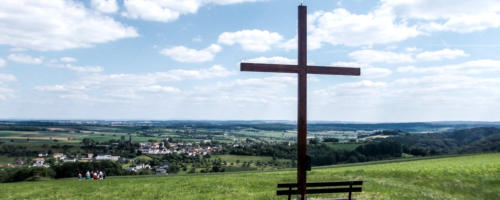 Holzkreuz auf einer Wiese mit Bank, im Hintergrund eine weite Landschaft und ein Dorf unter blauem Himmel mit Wolken., &copy; TI Bitburger Land
