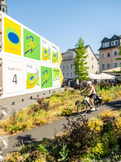 A signpost with bicycle symbols indicates the direction. In the background, a cyclist passes by, surrounded by a green area and buildings.