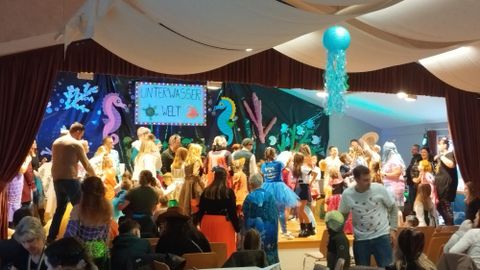 A lively carnival celebration with many costumed participants on a stage. Colorful decorations and a cheerful atmosphere characterize the scene.