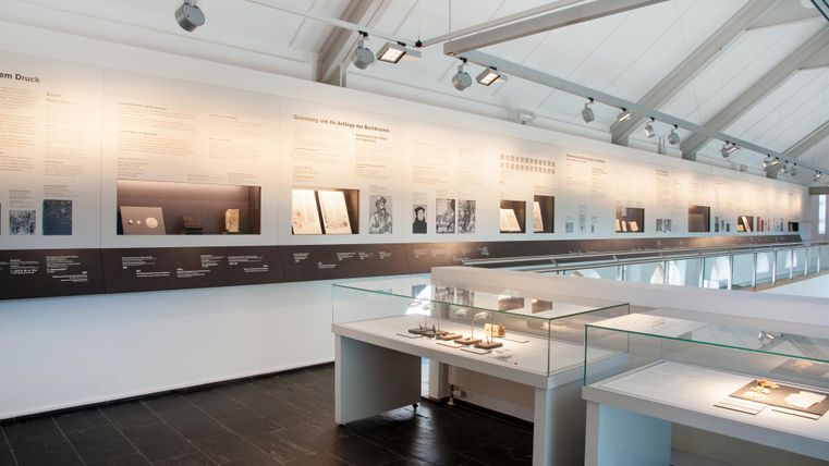 Ein modernes Museum mit einer Wand voller Informationen und Exponaten. Im Vordergrund sind Vitrinen mit verschiedenen Objekten zu sehen.