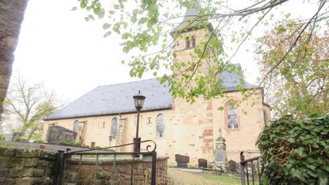 Een oude kerk met een hoge toren is omringd door bomen. De lucht is grijs en bewolkt.