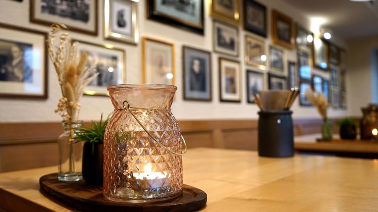 A cozy table with a candle in a glass and decorative plants. In the background, many framed pictures hang on the wall.