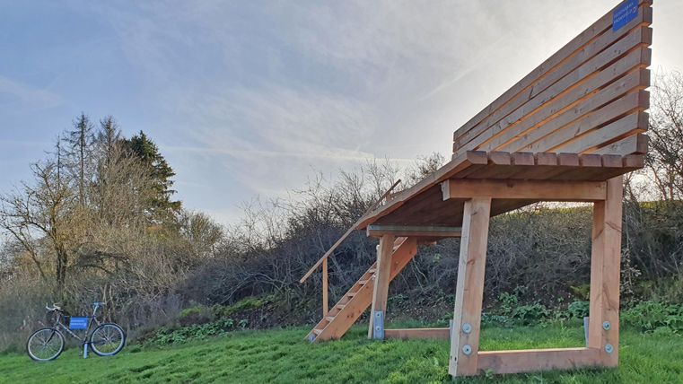 Un grand banc en bois se trouve sur une prairie, à côté d'un vélo. À l'arrière-plan, on voit des arbres et un ciel clair.