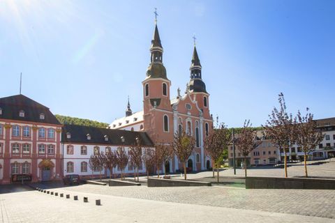 A historic building with two towers stands in a spacious square. Surrounded by trees and under a clear blue sky.