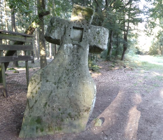 Une croix de pierre, connue sous le nom de croix de Fraubill, se dresse dans la for&ecirc;t. Elle est entour&eacute;e d'arbres et un chemin traverse la clairi&egrave;re., &copy; Elke Wagner, Felsenland S&uuml;deifel Tourismus GmbH