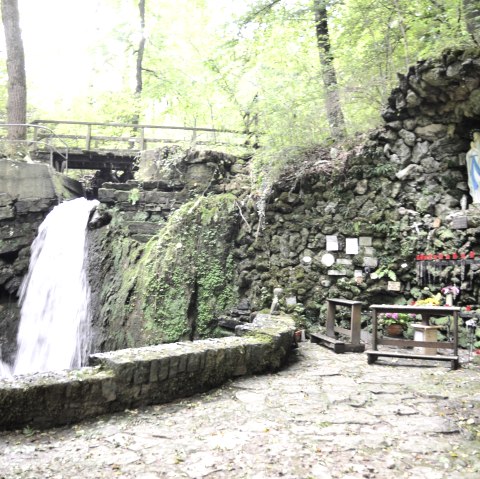 Die Mariengrotte Albach zeigt zwei Wasserf&auml;lle, eine Marienstatue in einer Felsnische und Kerzen auf einem Altar. Umgeben von &uuml;ppigem Gr&uuml;n., &copy; TI Bitburger Land