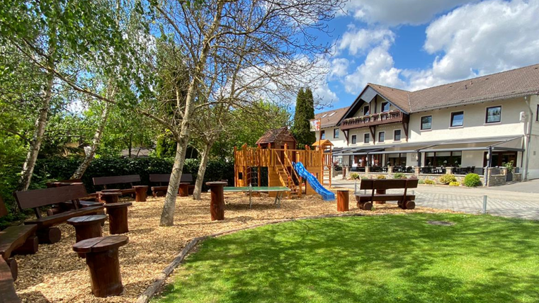 A beautiful outdoor area with a play corner, wooden seats, and a slide. In the background, there is a cozy building with a terrace.