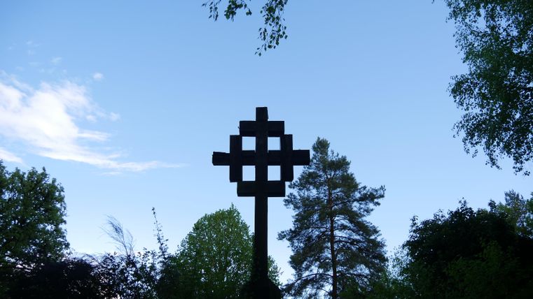 A large black cross stands in the foreground against a clear blue sky. Surrounded by green trees, the scene conveys a serene atmosphere.
