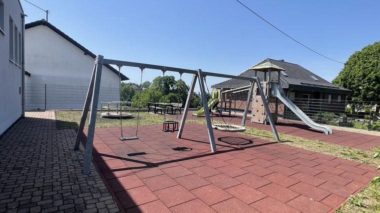 A playground with a swing and a slide. The ground is covered with red rubber tiles and there is plenty of grass areas.
