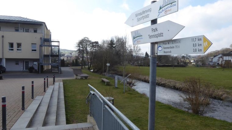 Een wegwijzer geeft verschillende bestemmingen aan, waaronder een park en het stadsplein. Op de achtergrond zijn een gebouw en een groen grasveld te zien.