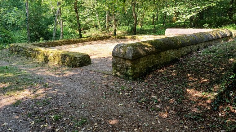 Een oude stenen structuur in een bosrijk gebied, omgeven door bomen en gevallen bladeren. Het pad leidt langs de historische ruïne.