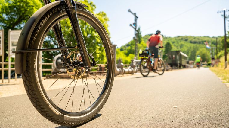 Ein Fahrradrad steht im Vordergrund auf einer sonnigen Straße. Im Hintergrund fahren zwei Radfahrer am Waldrand vorbei.
