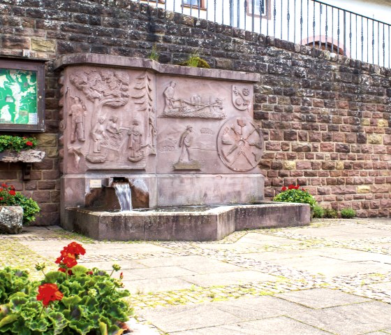 Une fontaine de village en pierre avec des reliefs et de l'eau qui coule, entour&eacute;e de fleurs rouges et d'un panneau d'information sur un mur en pierre., &copy; TI Bitburger Land - M. Mayer