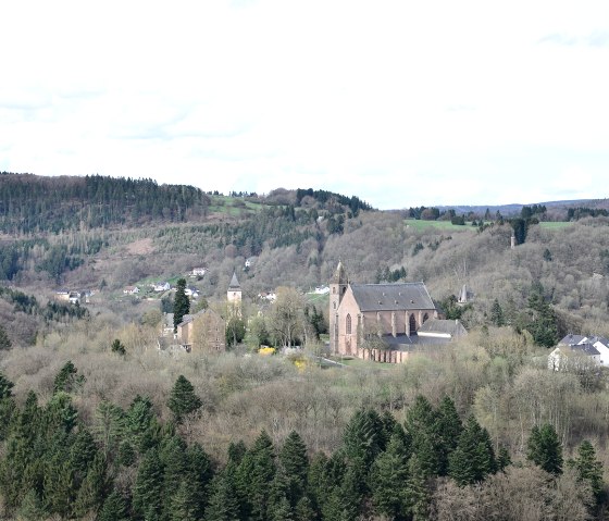 Gezicht op een kerk midden in een bosrijk landschap met heuvels op de achtergrond, genomen vanaf de Wilsecker Linde., &copy; TI Bitburger Land