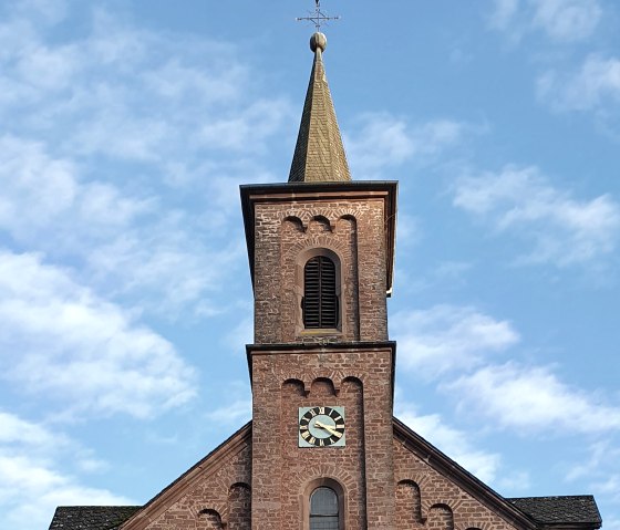 Backsteinkirche St. Laurentius mit Turm und Uhr, umgeben von B&auml;umen und einem Kreuz im Vordergrund, unter blauem Himmel., &copy; TI Bitburger Land