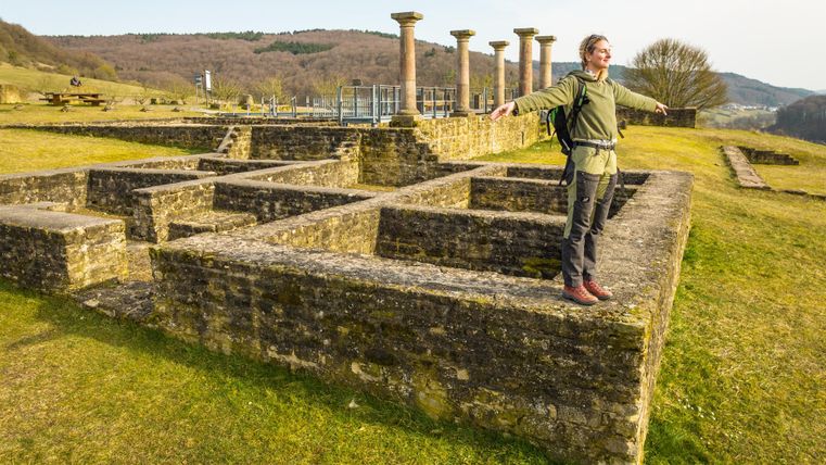 Ein Mann steht auf alten Ruinen in einer weitläufigen, grünen Landschaft. Im Hintergrund sind Säulen und sanfte Hügel zu sehen.