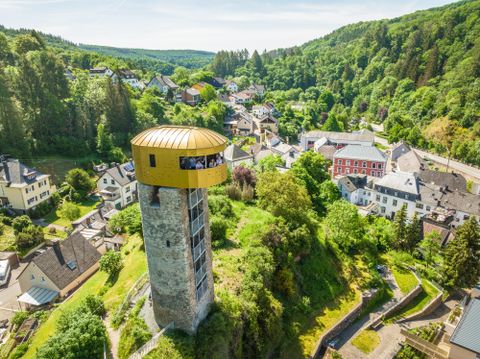 Een historische toren met een modern, gouden dak staat boven een schilderachtig stadje. Op de achtergrond strekken groene heuvels en bossen zich uit.