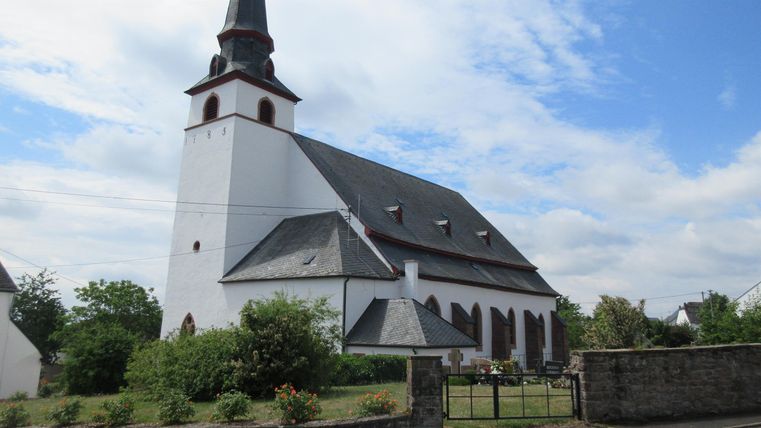 Eine weiße Kirched mit einem hohen Turm und schwarzem Dach. Umgeben von Bäumen und einem blauen Himmel.