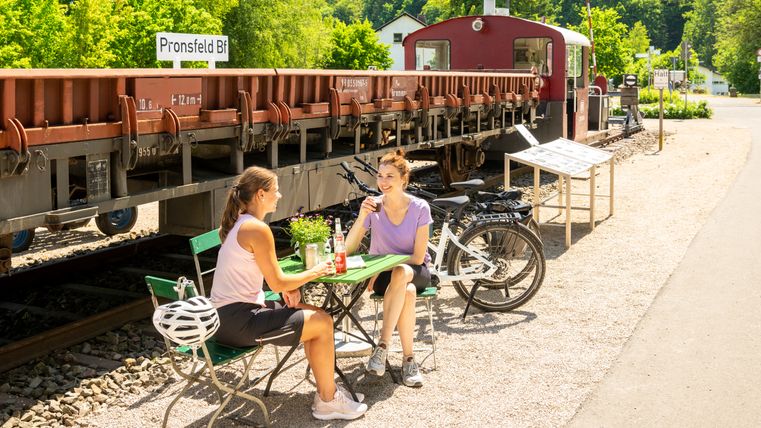 Zwei Frauen sitzen an einem Tisch im Freien vor einem alten Zug im Eisenbahnmuseum Pronsfeld.