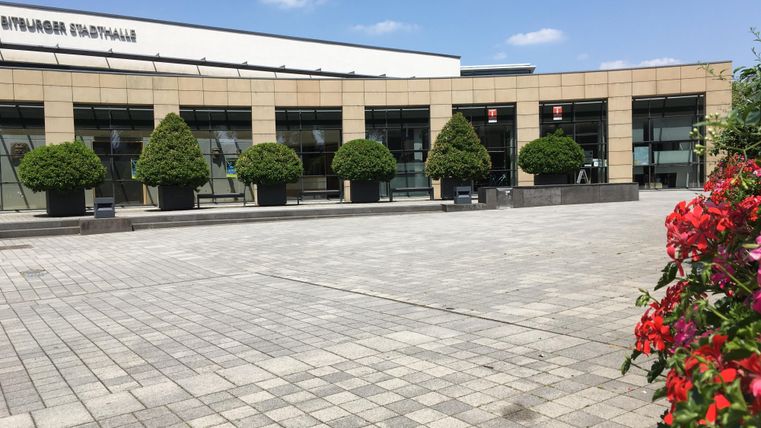 Ein moderner Gebäudekomplex mit großen Fenstern und einem gepflasterten Platz. Im Vordergrund sind blühende Pflanzen zu sehen.