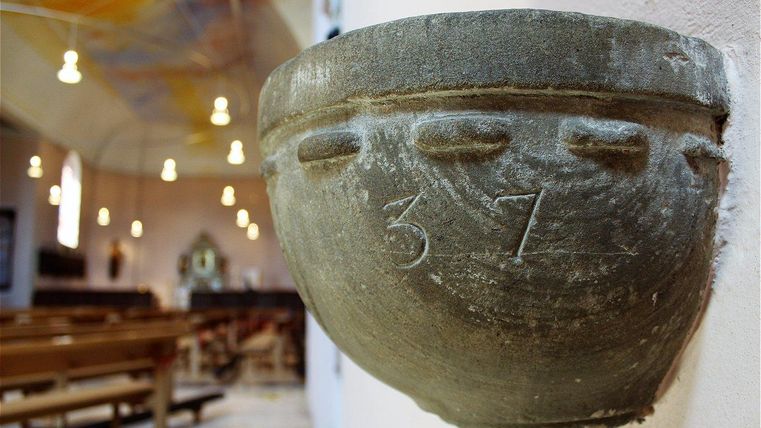 Eine steinerne Schale mit der Zahl 37 ist an der Wand einer Kirche befestigt. Im Hintergrund sind Kirchenbänke und sanfte Beleuchtung sichtbar.