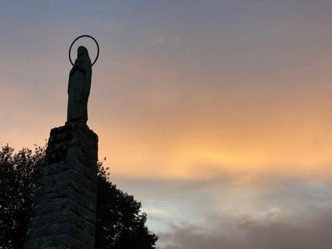 Une statue avec une auréole se dresse sur un haut socle. Le ciel est plongé dans des couleurs douces au coucher du soleil.