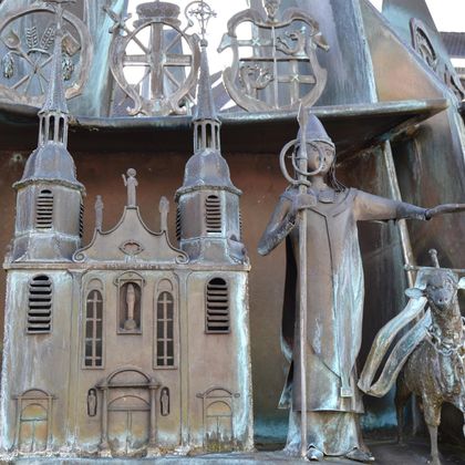 Une sculpture en métal artistique représente un évêque avec un bâton et une église. À l'arrière-plan, d'autres ornements sont visibles, rappelant des histoires ou des traditions.