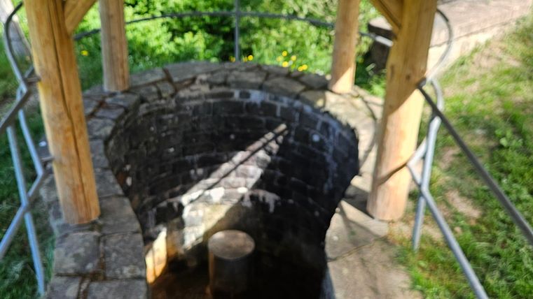 An old well with stone steps and a wooden overhang. Green surroundings and a sign are also visible.