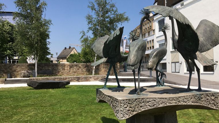 A modern sculpture of birds stands on a pedestal in a green park landscape. In the background, trees and buildings are visible.
