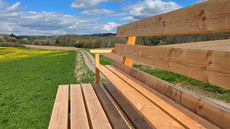 Un banc en bois est situé sur un champ vert sous un ciel bleu avec des nuages. À l'arrière-plan, on peut voir des prairies et des arbres.