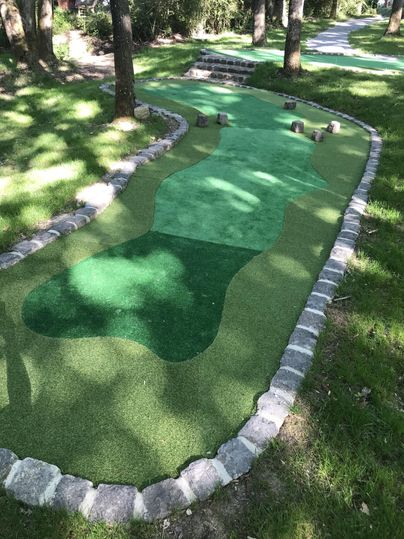 A mini golf course with green artificial grass and stone borders. The facility is surrounded by trees and features various hills and curves.