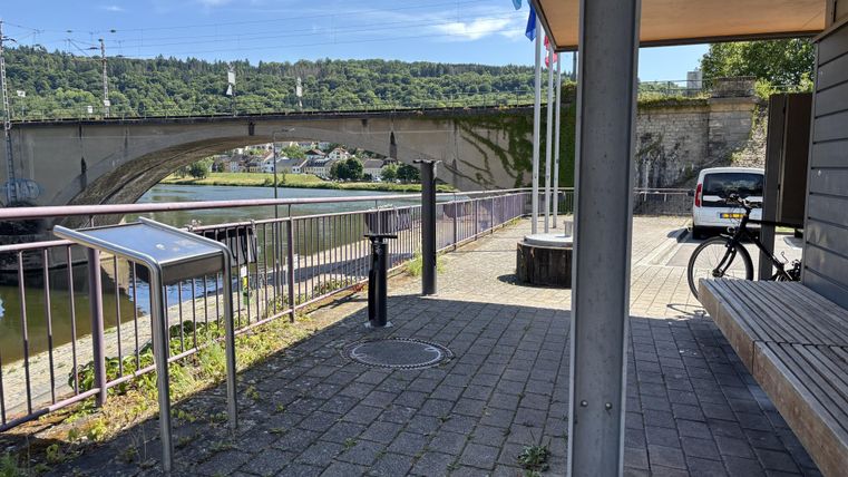 Ein ruhiger Uferbereich mit einer Brücke im Hintergrund. Auf dem Platz stehen Fahrräder und eine Sitzgelegenheit.