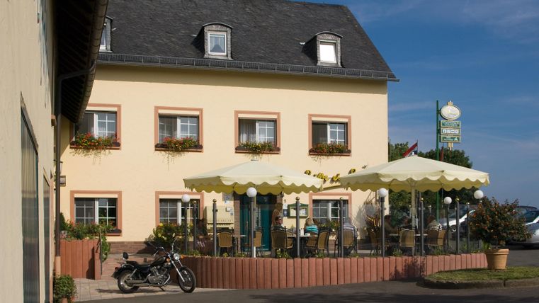 Ein einladendes Gasthaus mit hellem Fassaden und einer Terrasse, die von Sonnenschirmen geschützt ist. Vor dem Gebäude steht ein Motorrad, und im Hintergrund ist ein klarer Himmel zu sehen.