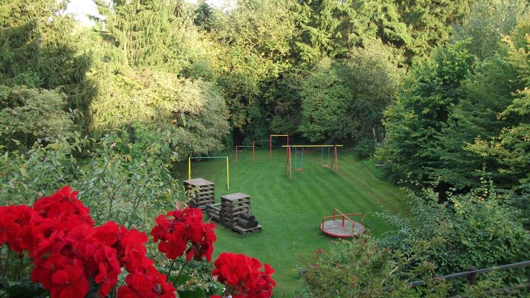 A beautiful garden with lots of greenery and trees. In the foreground, red flowers are blooming, while in the background, swings and a wooden platform can be seen.