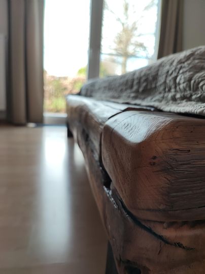 A rustic wooden sofa with a natural surface and clear lines. In the background, there are windows with daylight and a green view.