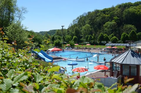 A swimming pool with a blue slide and many colorful beach umbrellas. In the background, there are green trees and many people enjoying the water.