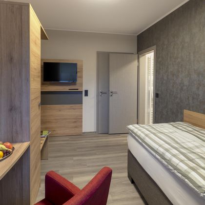 Ein modernes Hotelzimmer mit einem Einzelbett, einem Fernseher an der Wand und einem roten Stuhl. Die Wände sind in neutralen Farben gehalten und der Boden ist aus Holz.