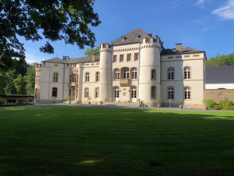 Ein elegantes Schloss mit Türmen und großen Fenstern, umgeben von grünem Rasen. Der Himmel ist blau mit wenigen Wolken.