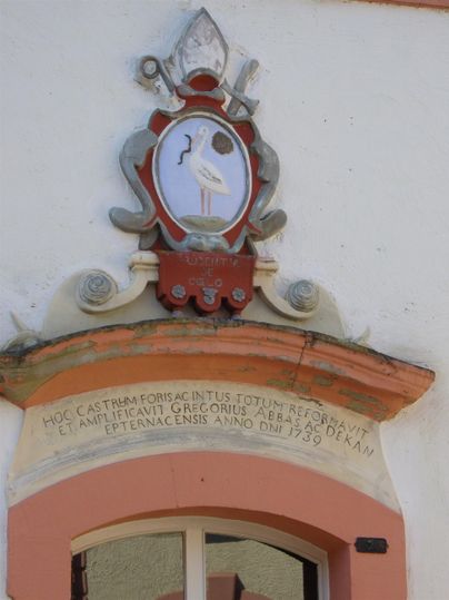 A coat of arms with a stork and a medallion-like background is affixed to a building wall. Below the coat of arms is an inscription in cursive script.