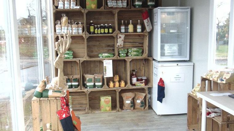 Ein kleiner Verkaufsraum mit Holzregalen, die mit verschiedenen Produkten gefüllt sind. Auf der rechten Seite steht ein Kühlschrank und ein Tisch mit weiteren Artikeln.