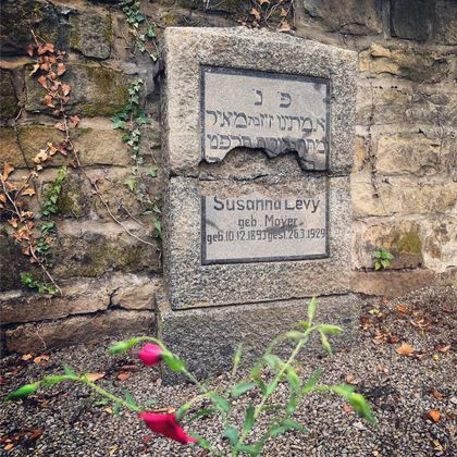 Ein Grabstein mit der Inschrift „Susanna Levy“ in einem ruhigen, steinernen Umfeld. Eine kleine Blume wächst in der Nähe des Grabes.