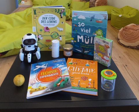 On a table lie various books about science and technology, a small robot, and a glass with colorful sections. There is also a lemon and a few other items that indicate learning and experimenting.