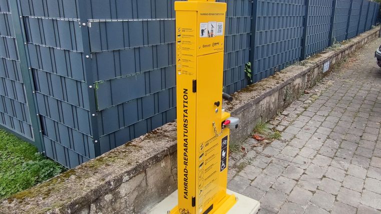 Een gele hydrantenpaal staat aan een trottoir. Op de achtergrond zijn grijze, moderne hekelementen zichtbaar.