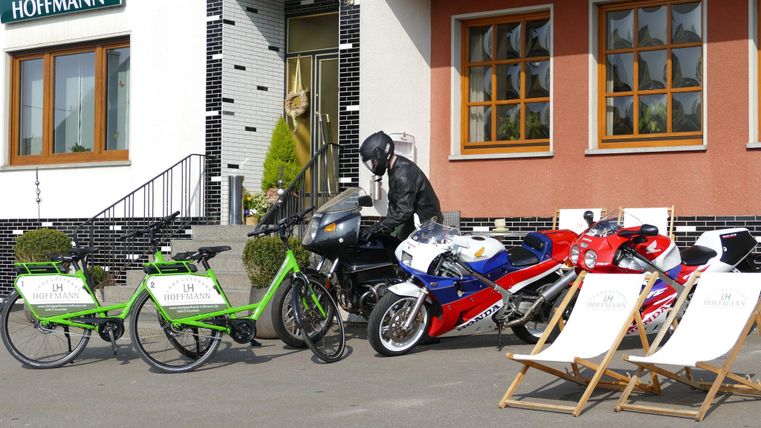 A row of bicycles and motorcycles is in front of a building. A motorcyclist in protective gear is standing next to the vehicles.