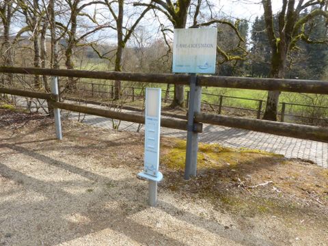 A signboard stands by a path with a scale next to it. The area is surrounded by trees and has a clear view of the landscape.