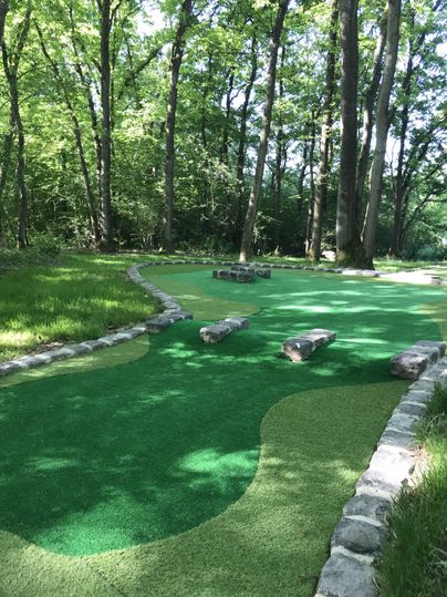 A mini-golf course in the woods with green artificial grass and stone obstacles. Surrounded by tall trees, it offers a pleasant natural ambiance.