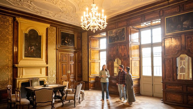 Ein elegantes Zimmer mit Holzwänden, das mit Gemälden dekoriert ist. Drei Personen stehen um einen Tisch und unterhalten sich.